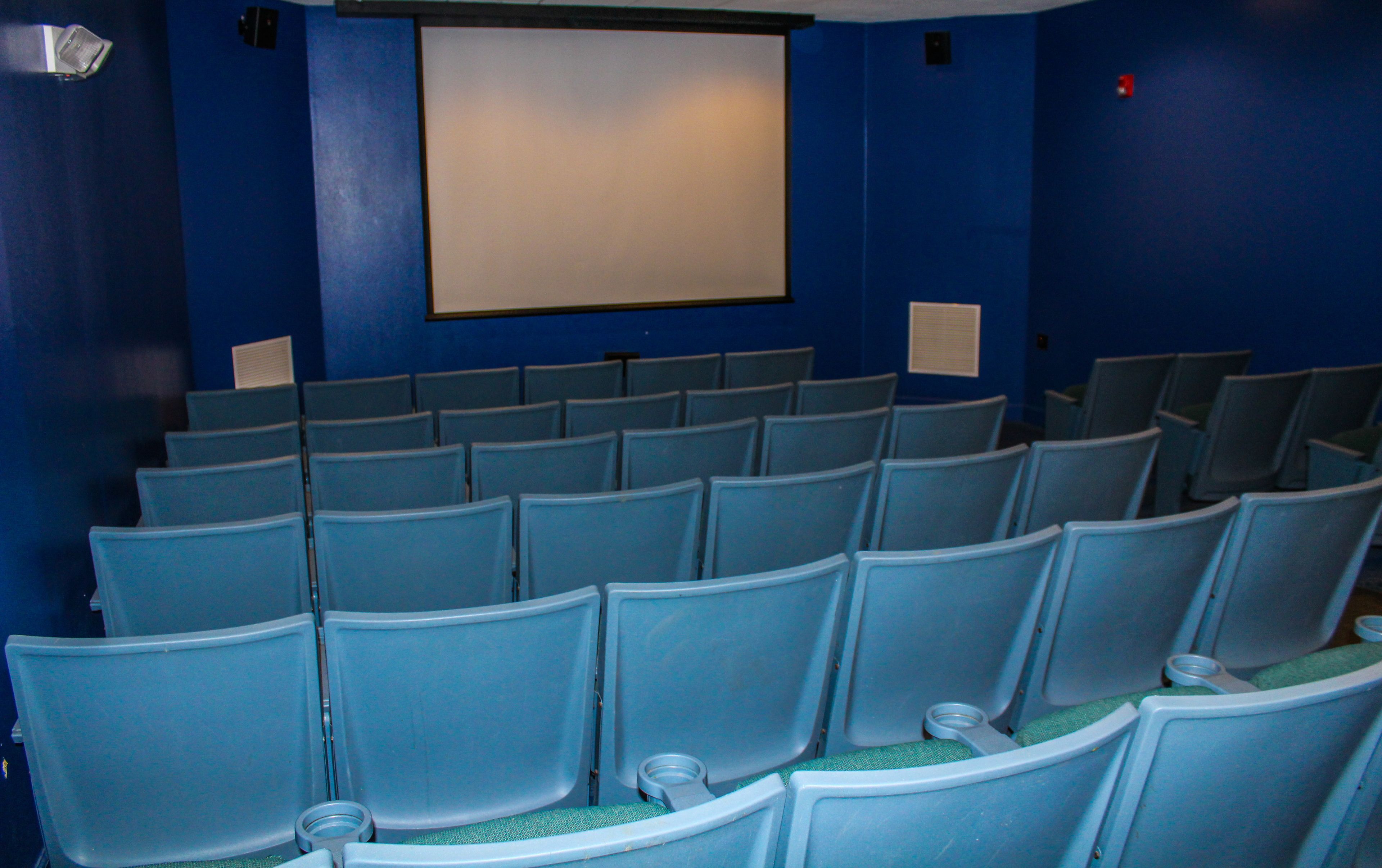 An indoor auditorium with rows of blue chairs, a large blank projection screen, and a blue wall.