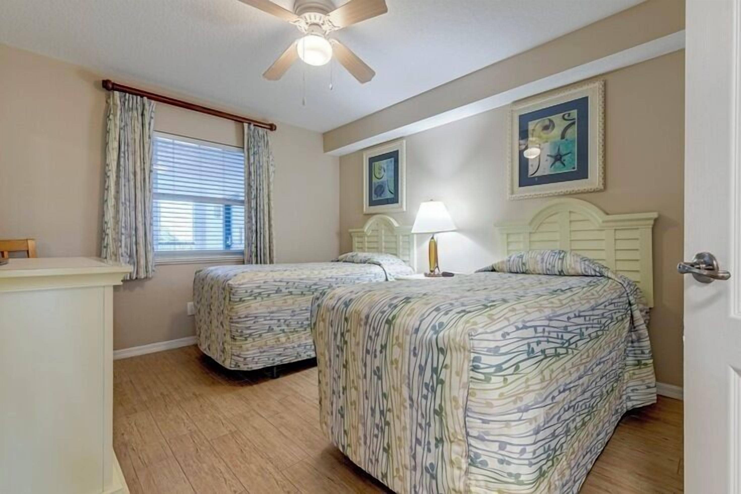 A bedroom with two beds, a ceiling fan, a window with curtains, and artwork on the walls.