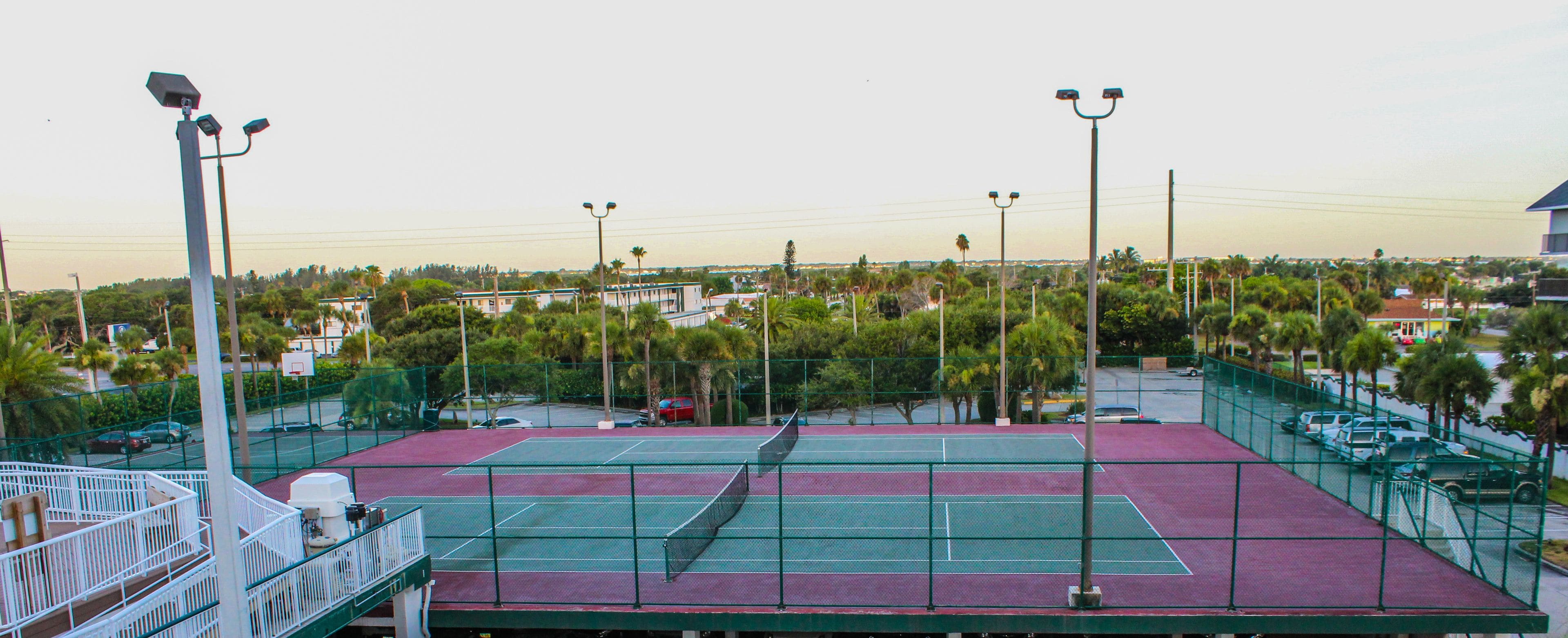A tennis court with a net, surrounded by a green fence, and streetlights in the background.