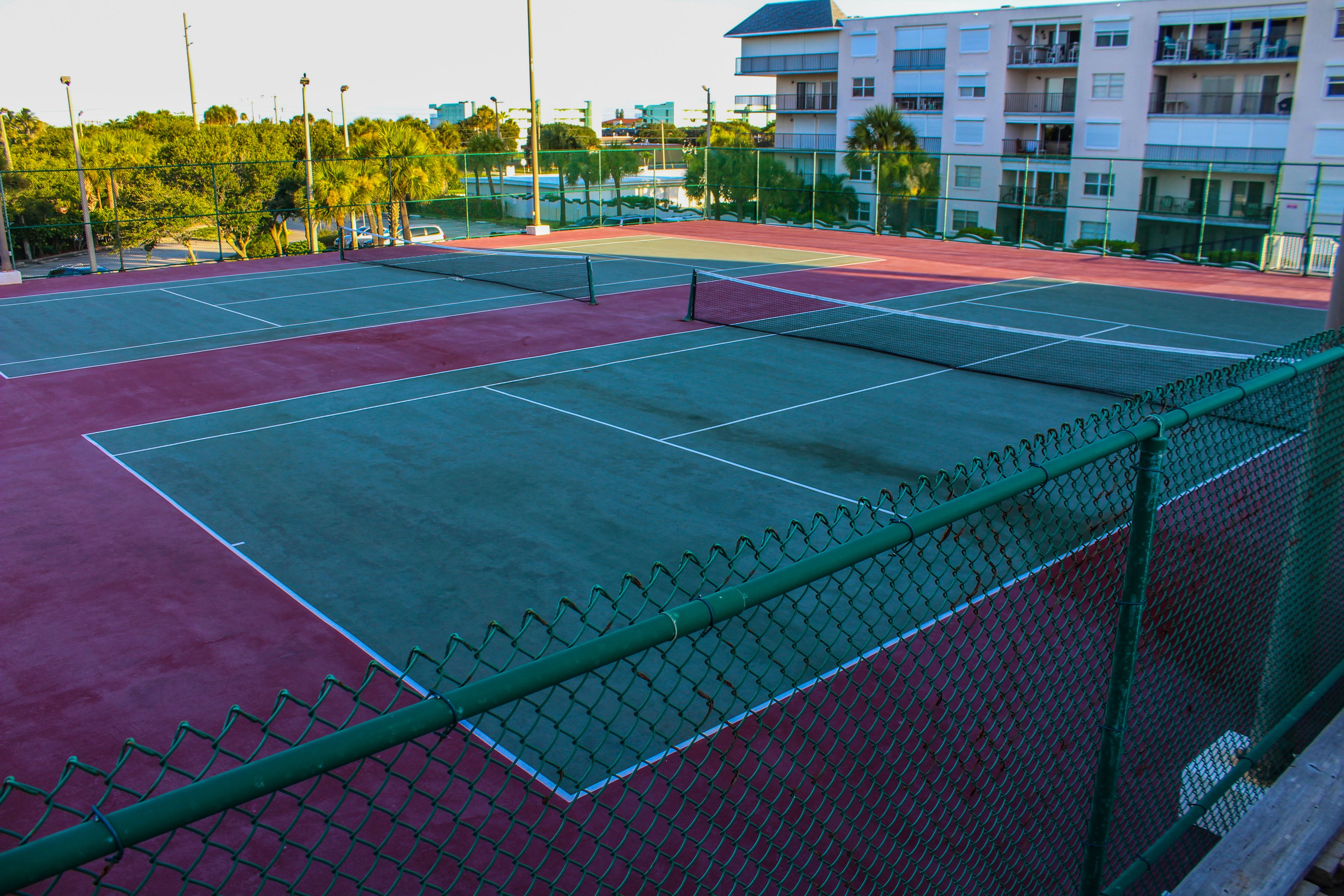 An outdoor tennis court with a green fence and a red surface.