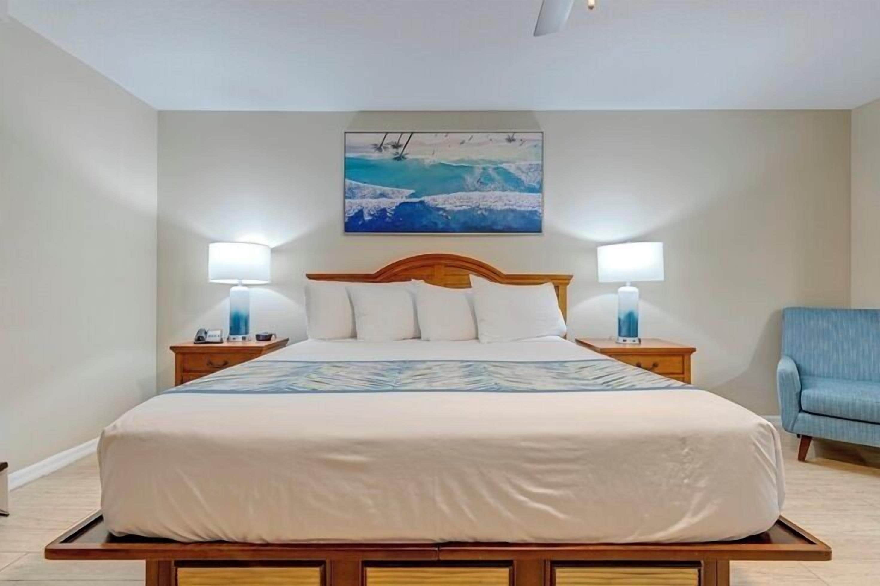 A neatly made bed with white linens and a wooden headboard, flanked by two bedside tables with lamps, in a room with a large painting on the wall.