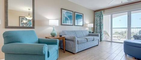 A spacious living room with a blue armchair, a grey sofa, and a balcony with a view.