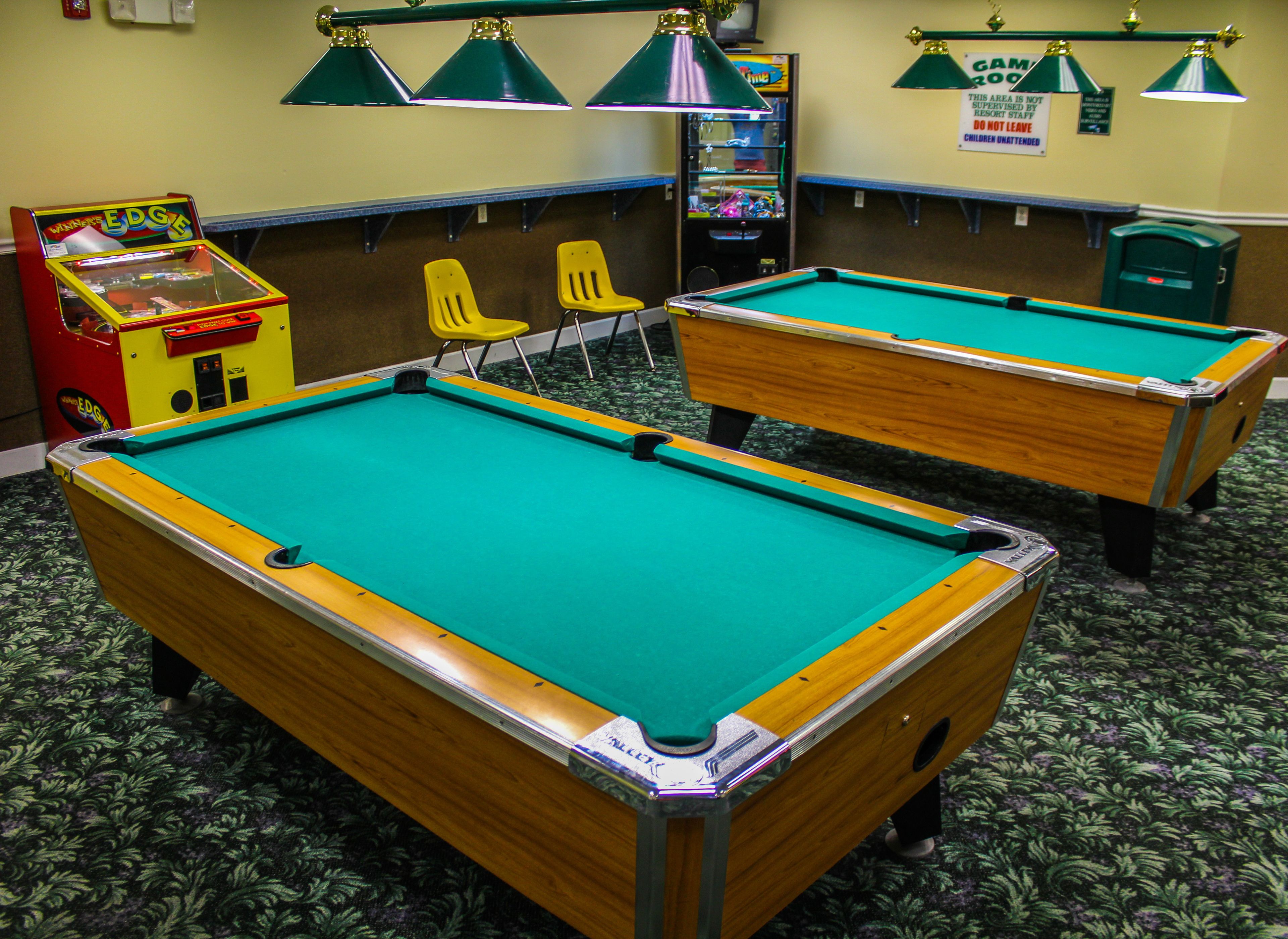 Deux tables de billard dans une pièce dont le sol est recouvert d’une moquette verte, avec des chaises jaunes, et une autre table de billard à l’arrière-plan.