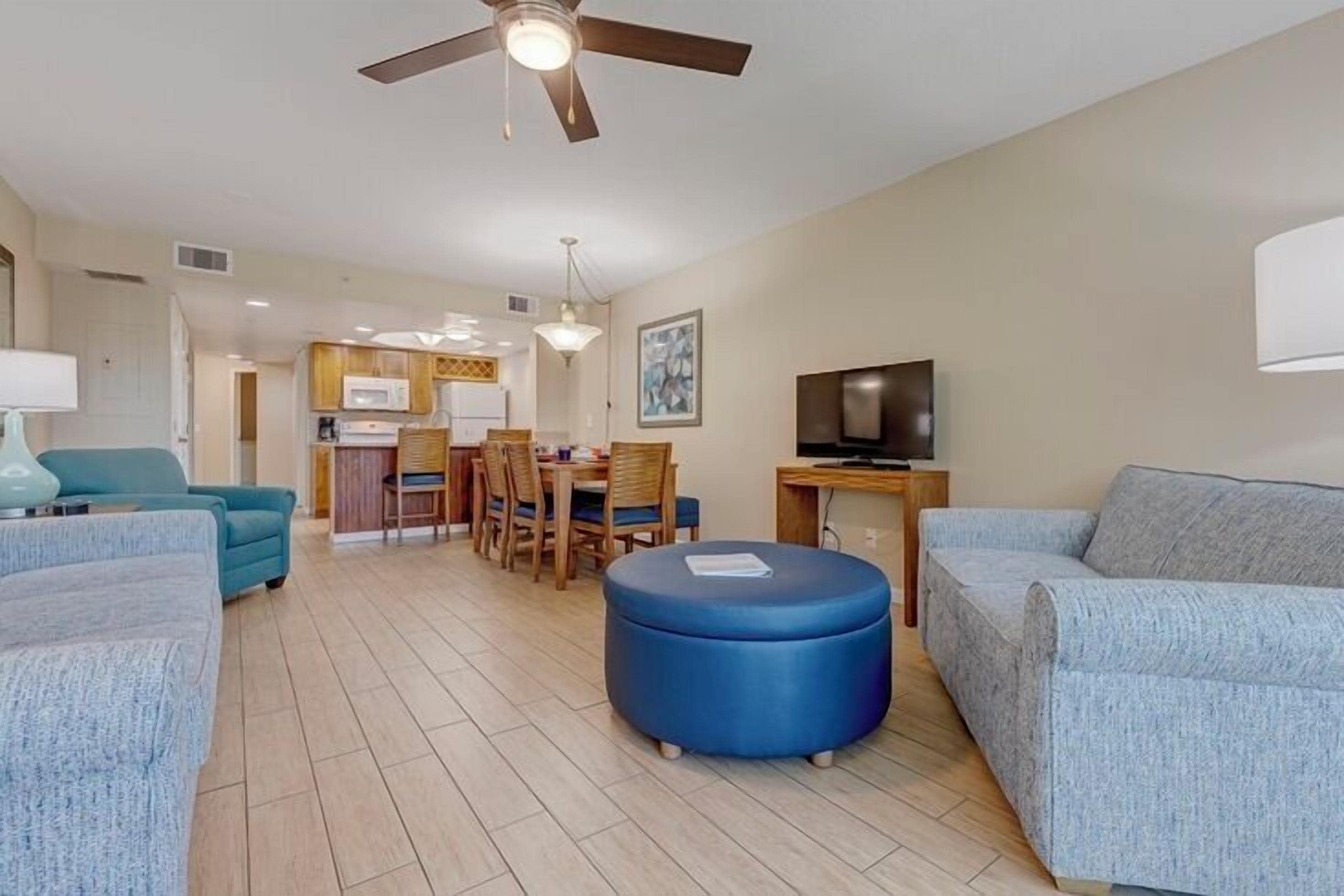 A living room with two sofas, a blue ottoman, a ceiling fan, a TV, and a kitchen area in the background.