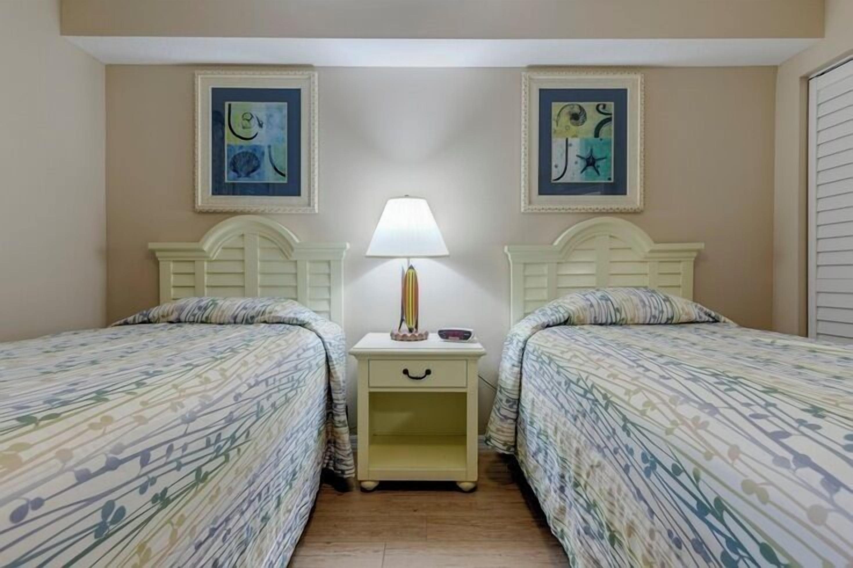Une chambre avec deux lits, chacun recouvert d’une couvre-lit à motifs, une table de chevet avec une lampe, et deux œuvres d’art encadrées accrochées au mur.