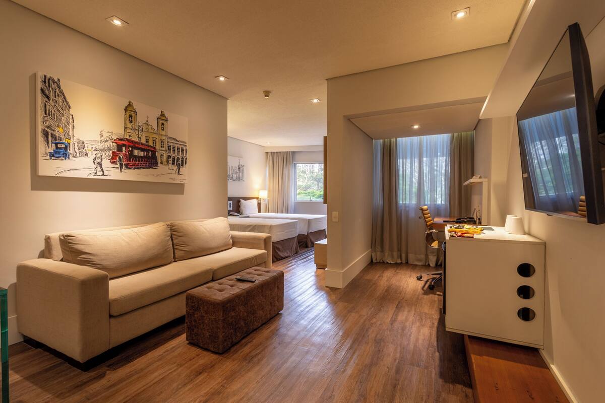 A modern hotel room with a sofa, ottoman, and a dining area with a table and chairs.