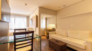 Quarto de hotel moderno com mesa de jantar de vidro, sofá branco, cadeira de madeira e um pequeno banquinho.
