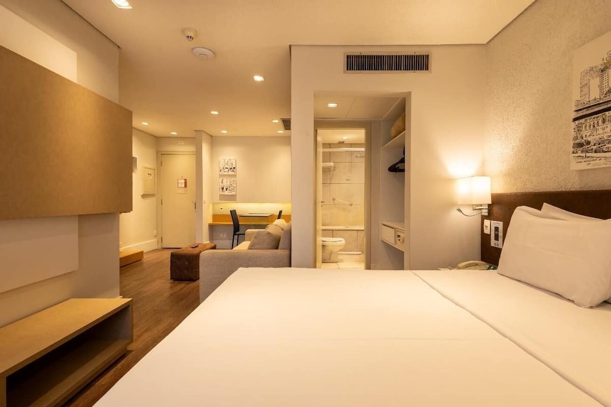 A hotel room with a bed, a desk, a chair, a bathroom, and a hallway.
