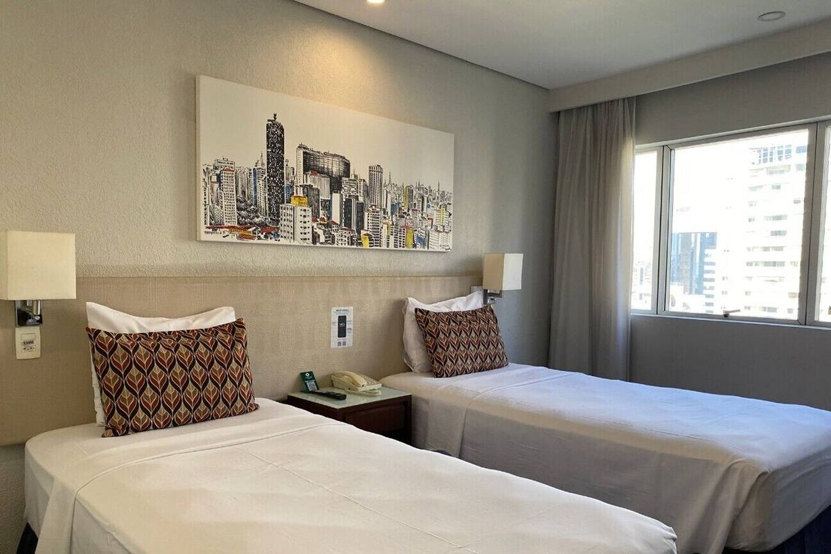 A hotel room with two beds, a cityscape painting, a window with curtains, and a bedside table with a phone.
