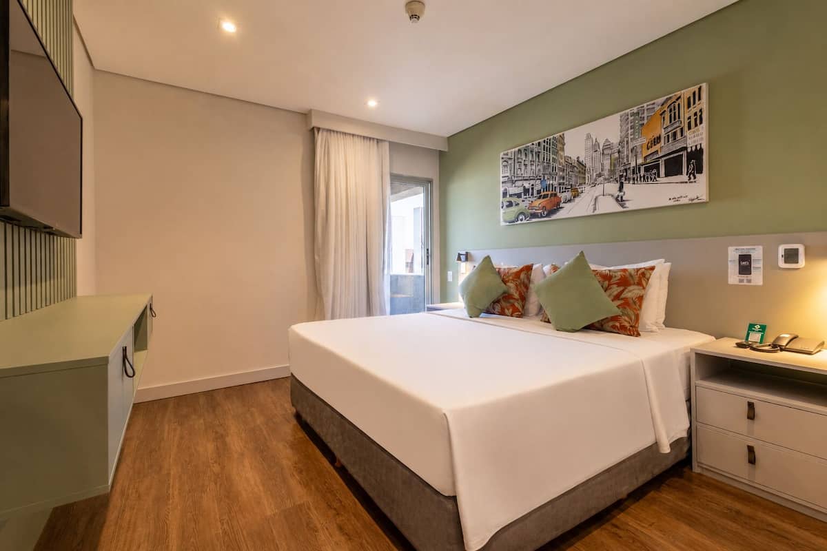A hotel room with a bed, a nightstand, a desk, a chair, a window with curtains, and a wall with a cityscape painting.