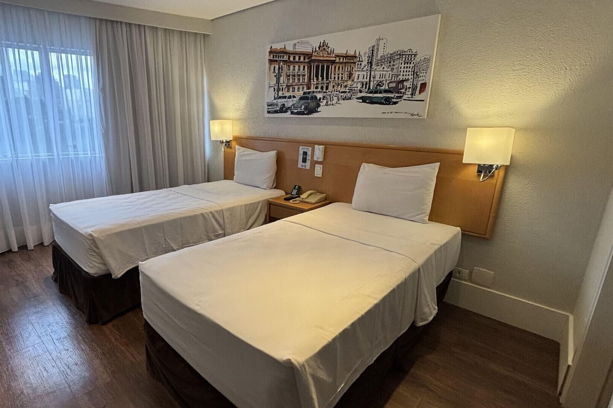 A hotel room with two beds, a window with curtains, a wall-mounted lamp, and a cityscape poster.