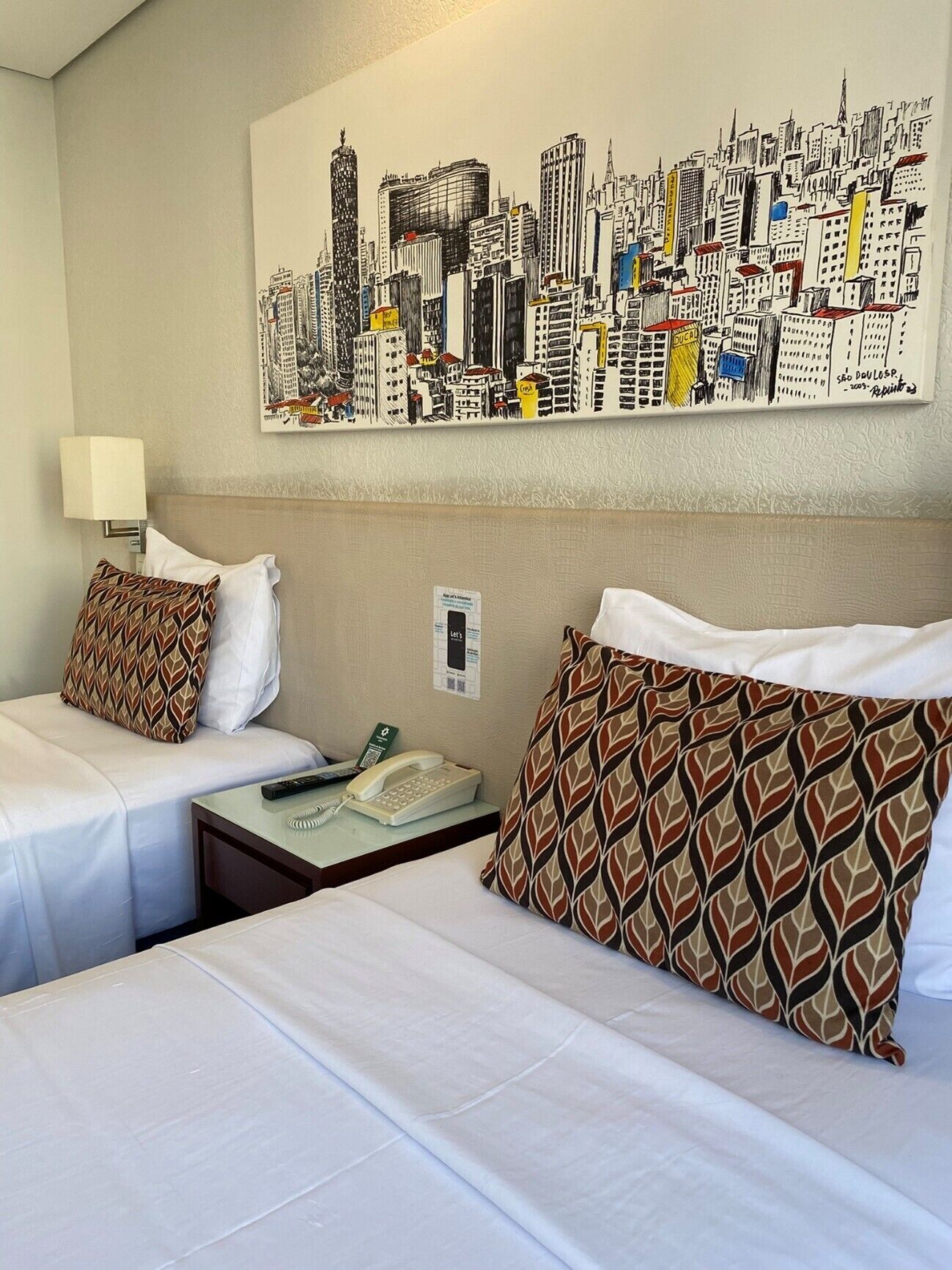 Habitación de hotel con dos camas, un cuadro con un paisaje urbano, una lámpara, un teléfono y una mesita.