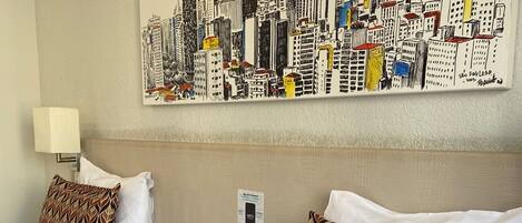 A hotel room with two beds, a cityscape artwork on the wall, a lamp, a phone, and a small table.