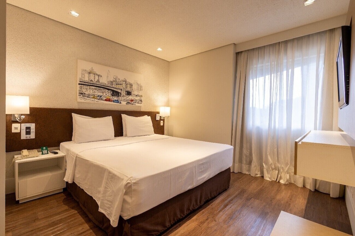 A hotel room with a large bed, two bedside tables with lamps, a wall-mounted TV, and a window with curtains.