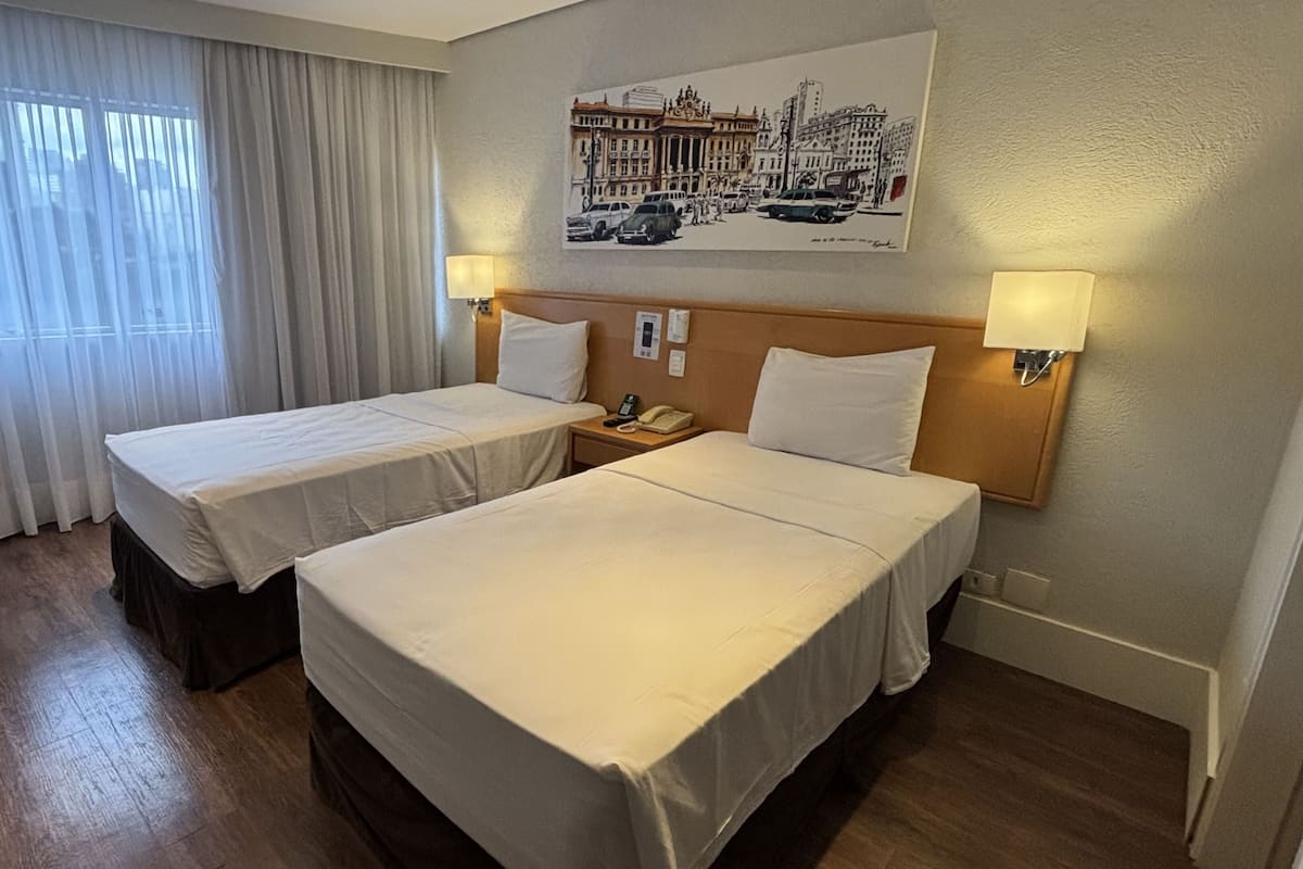 A hotel room with two beds, a window with curtains, a wall-mounted painting, and a nightstand with a lamp.