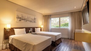 Habitación de hotel con dos camas, una obra de arte con paisaje urbano en la pared, un ventanal grande con cortinas y un televisor de pantalla plana en la pared.