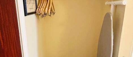 Pequeña habitación con puerta de madera, pared blanca, un certificado enmarcado, un tendedero con ropa, una bolsa negra y una tabla de planchar gris.