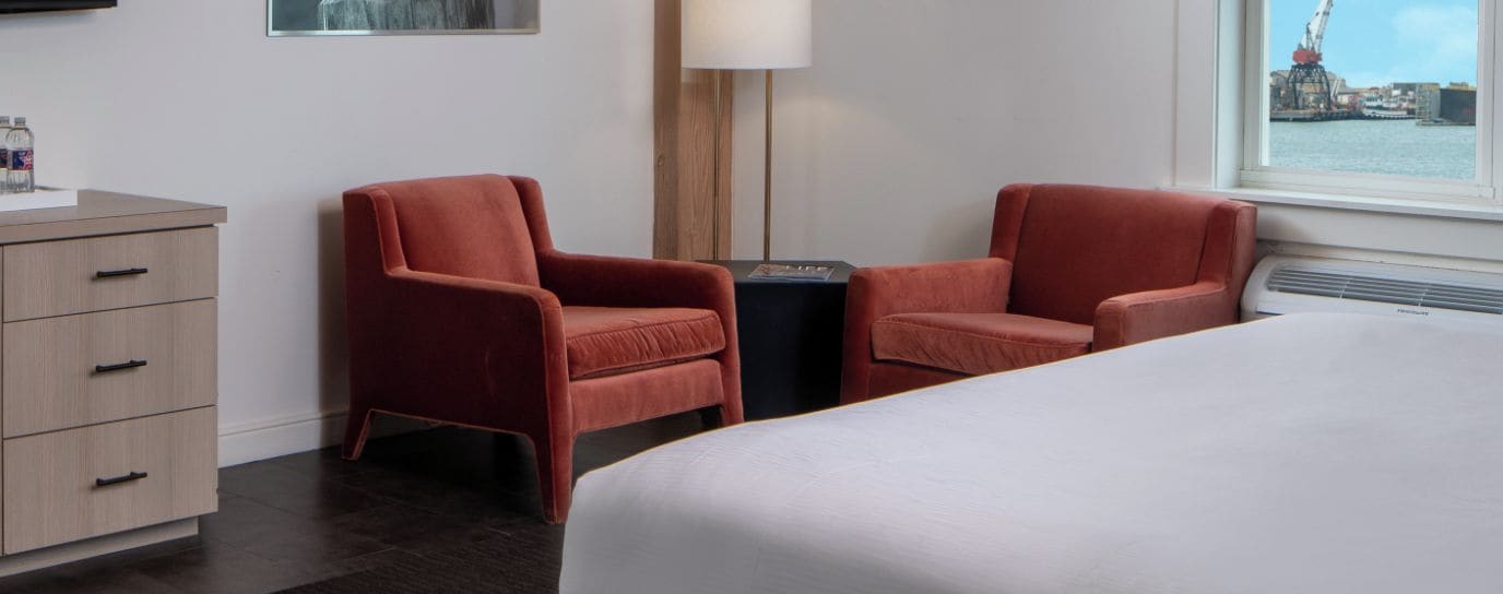 Habitación de hotel con una cama, dos sillones rojos, una mesita, una lámpara y vistas a un puerto con grúas.