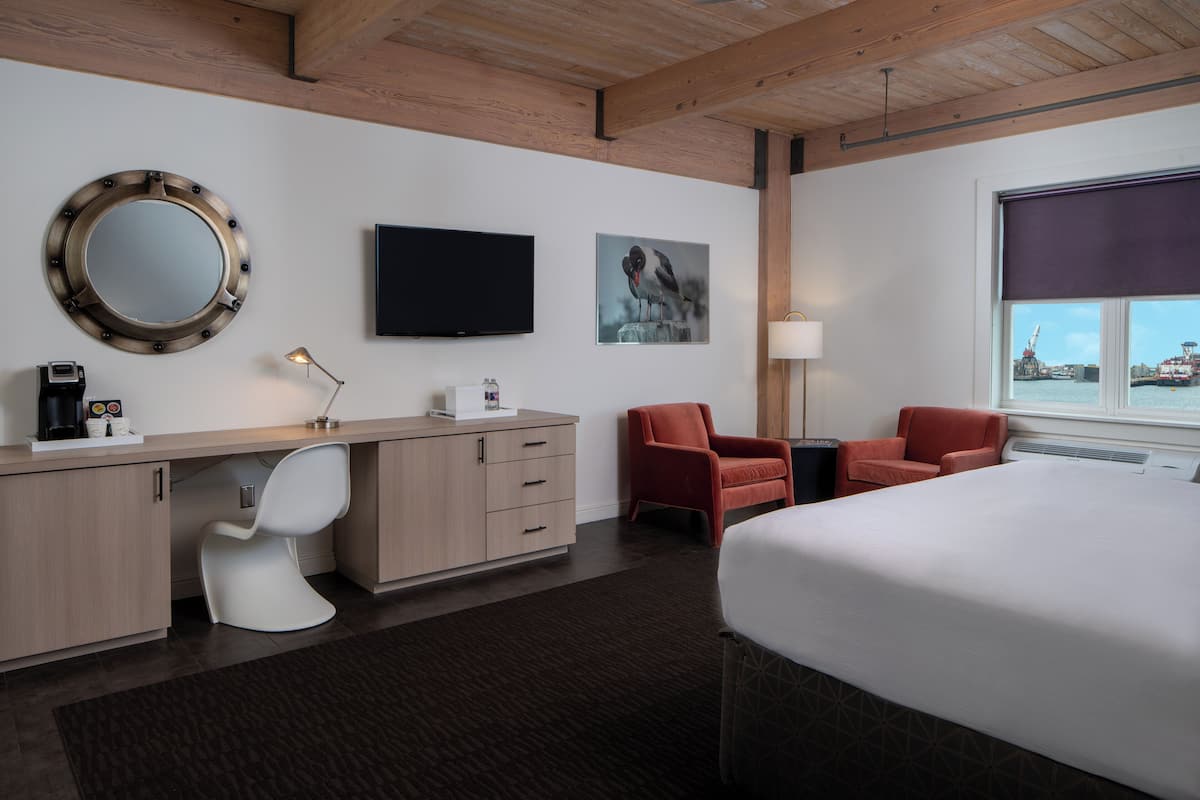 A modern hotel room with a wooden ceiling, a large bed, a desk with a chair, a television, and a window with a view of a crane.