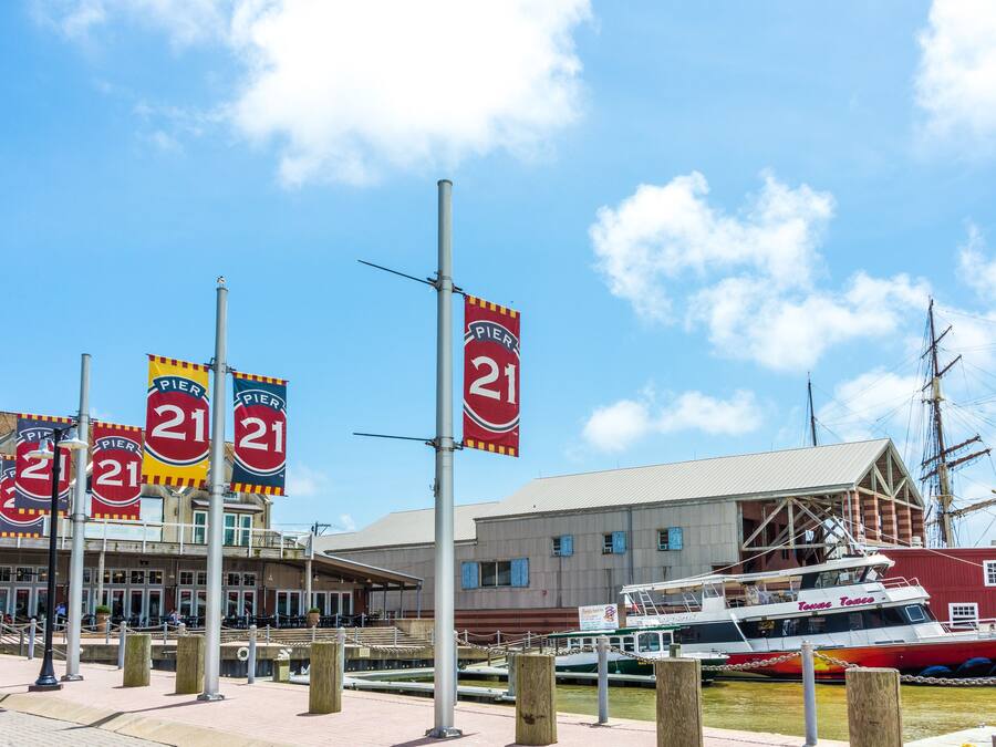 Un muelle con varios letreros que indican 'Pier 21', un edificio grande con techo plano y un barco histórico amarrado cerca.