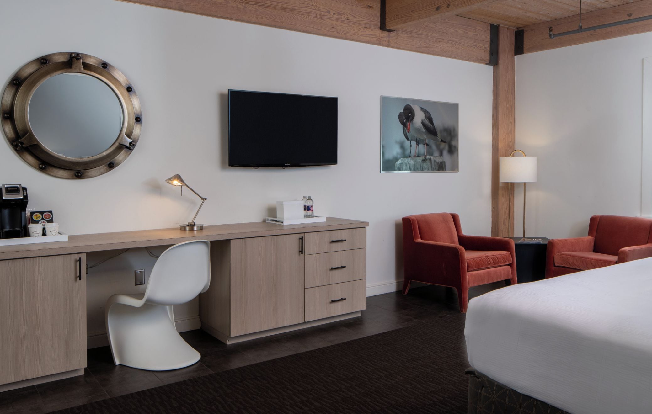 A hotel room with a desk, a chair, a bed, a TV, a painting, and a lamp.
