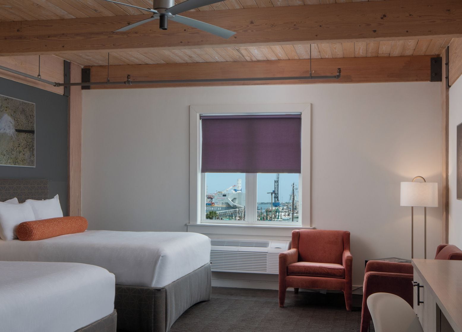 Ein Hotelzimmer mit zwei Betten, einem Sessel, einem Schreibtisch, einer Lampe und einem Fenster mit Blick auf ein Schiff.