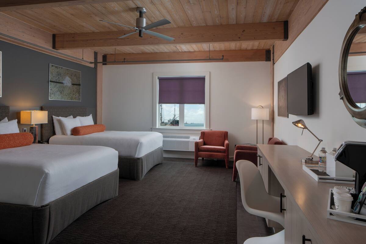A hotel room with two beds, a desk, a chair, a TV, a ceiling fan, a window with blinds, and a wall-mounted TV.