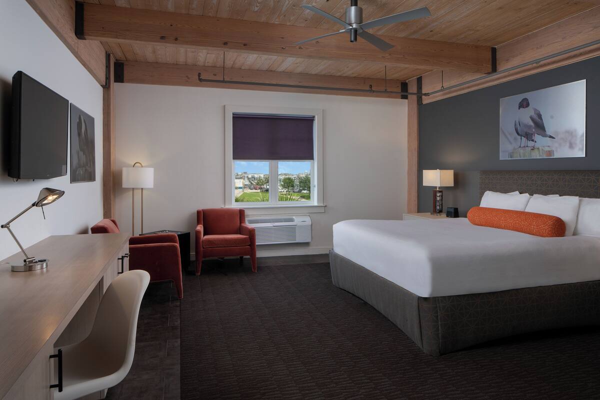 A hotel room with a bed, desk, chairs, a window with a view, and a ceiling fan.