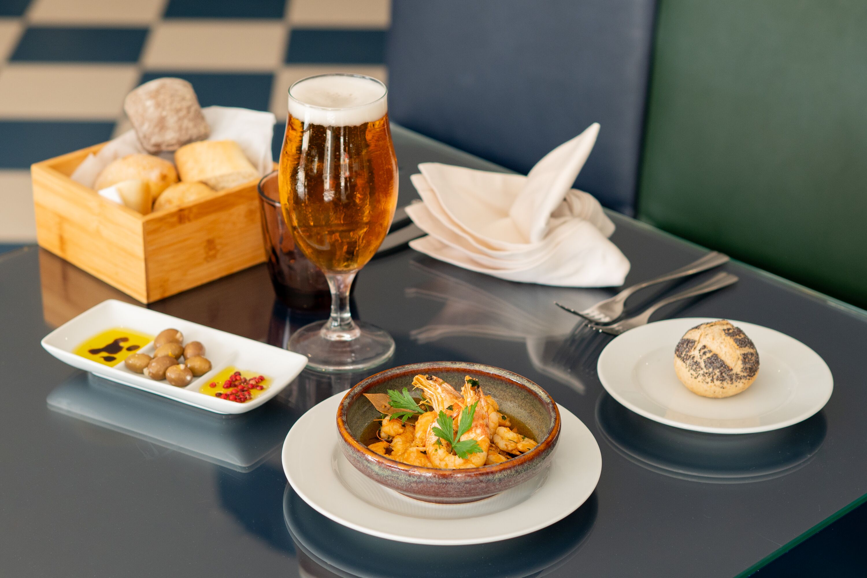 A dining table with a bowl of shrimp, a glass of beer, bread, and a small plate of olives and spices.