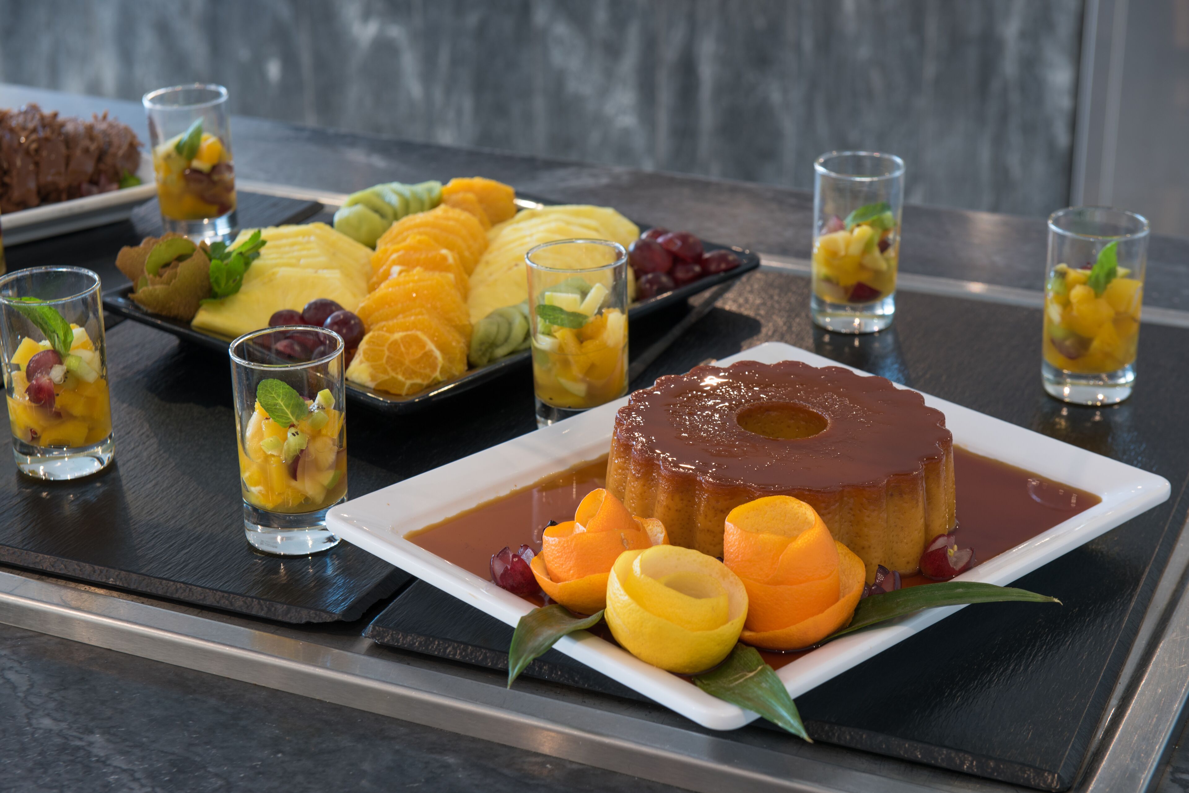 Un plato de postres con varias frutas y un flan de caramelo sobre una bandeja.