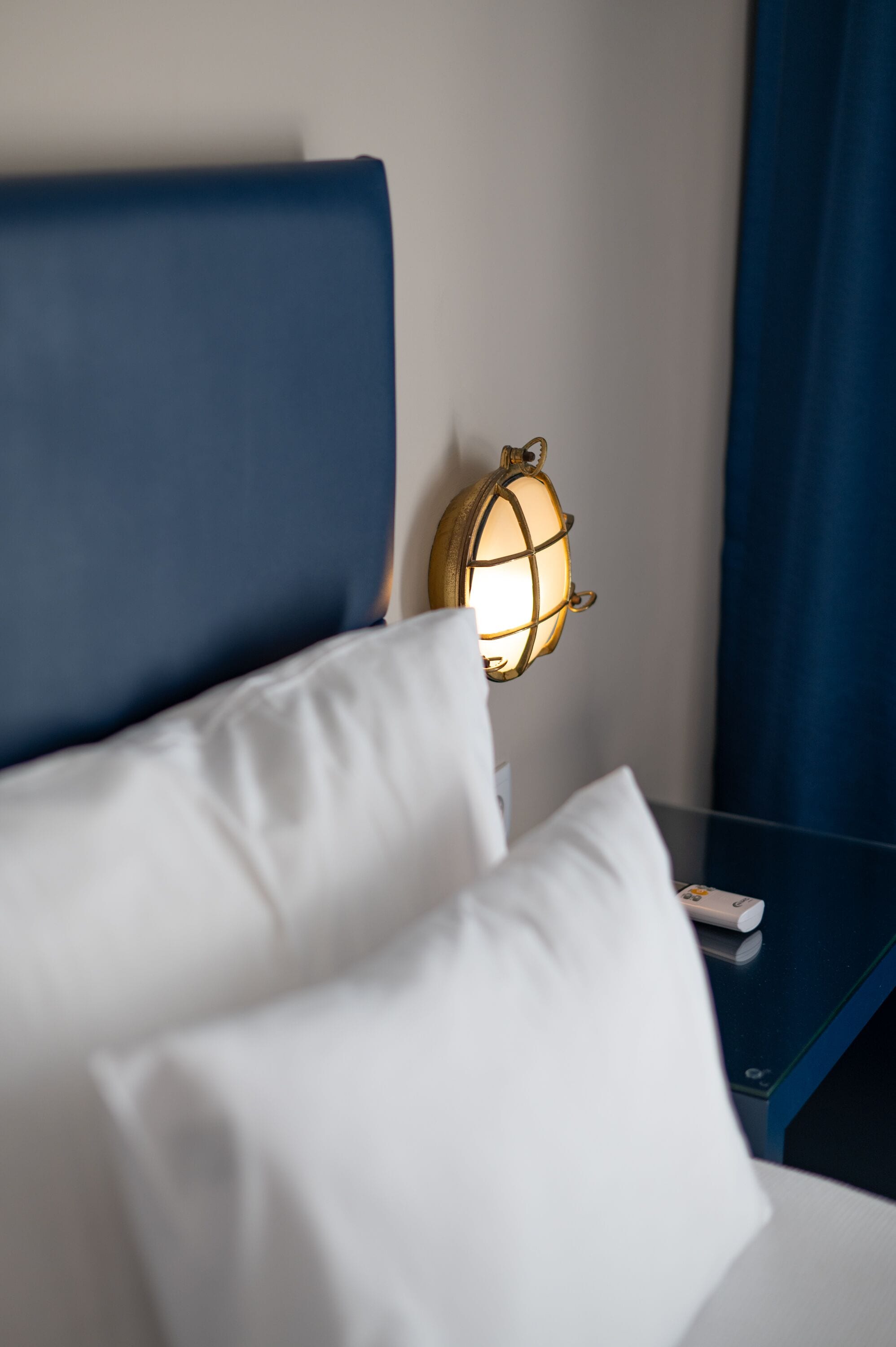 A neatly made bed with white pillows and a blue headboard. A small, round, illuminated wall lamp is visible on the wall beside the bed.