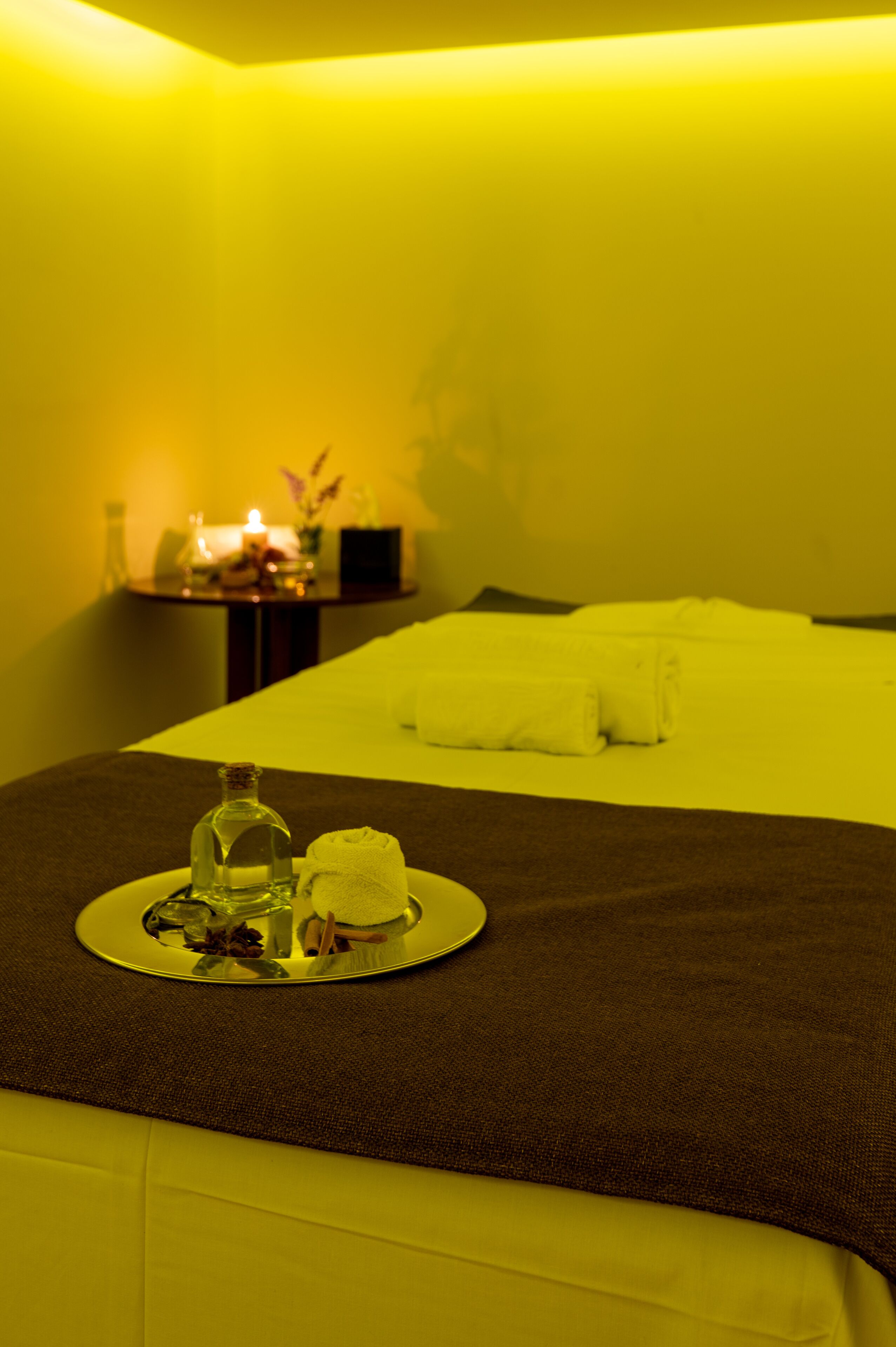 A hotel room with a bed, a tray with a bottle and a towel, and a nightstand with a candle and flowers.