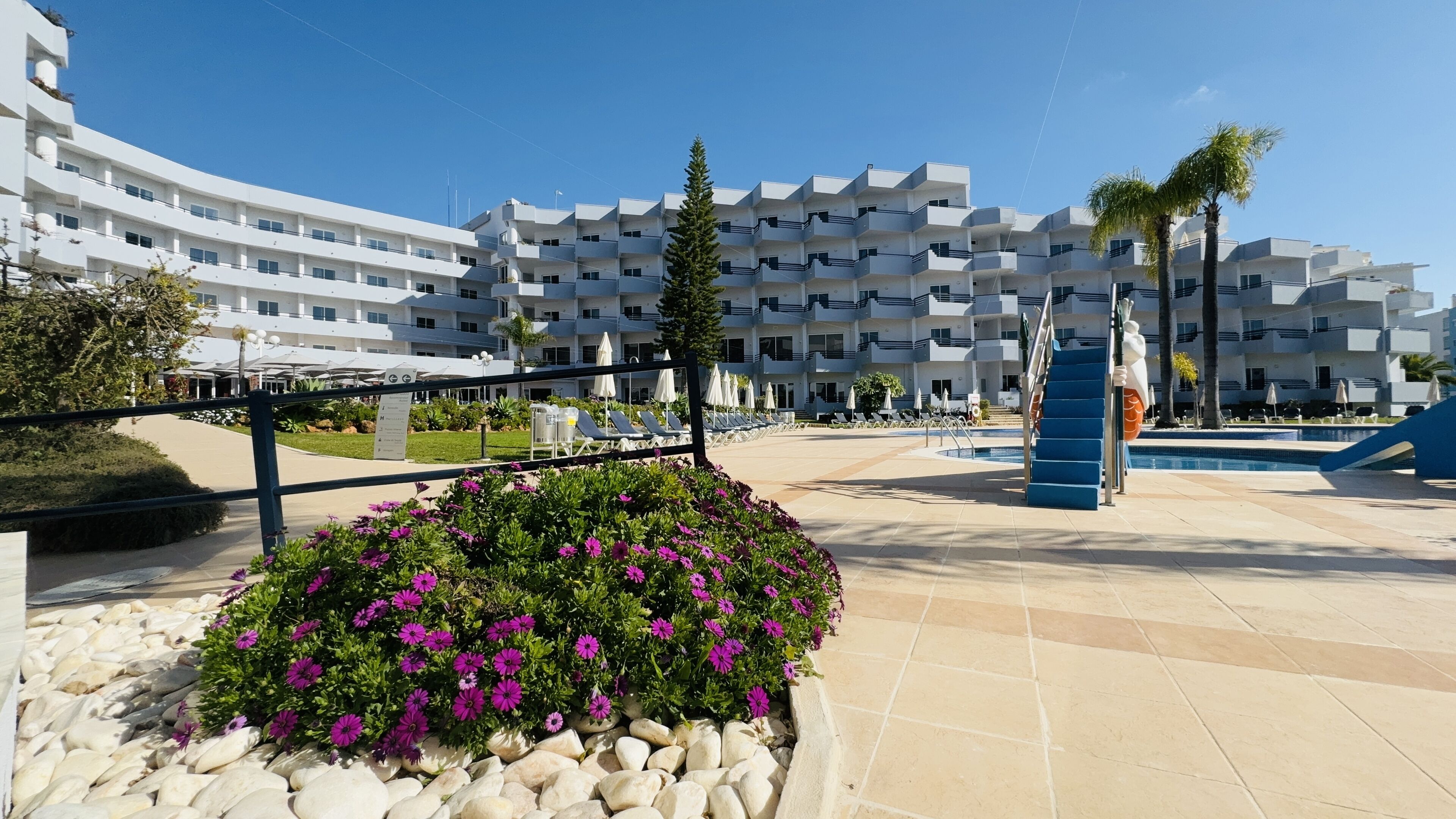 Um hotel moderno com várias varandas, uma piscina e um jardim ajardinado com flores roxas.