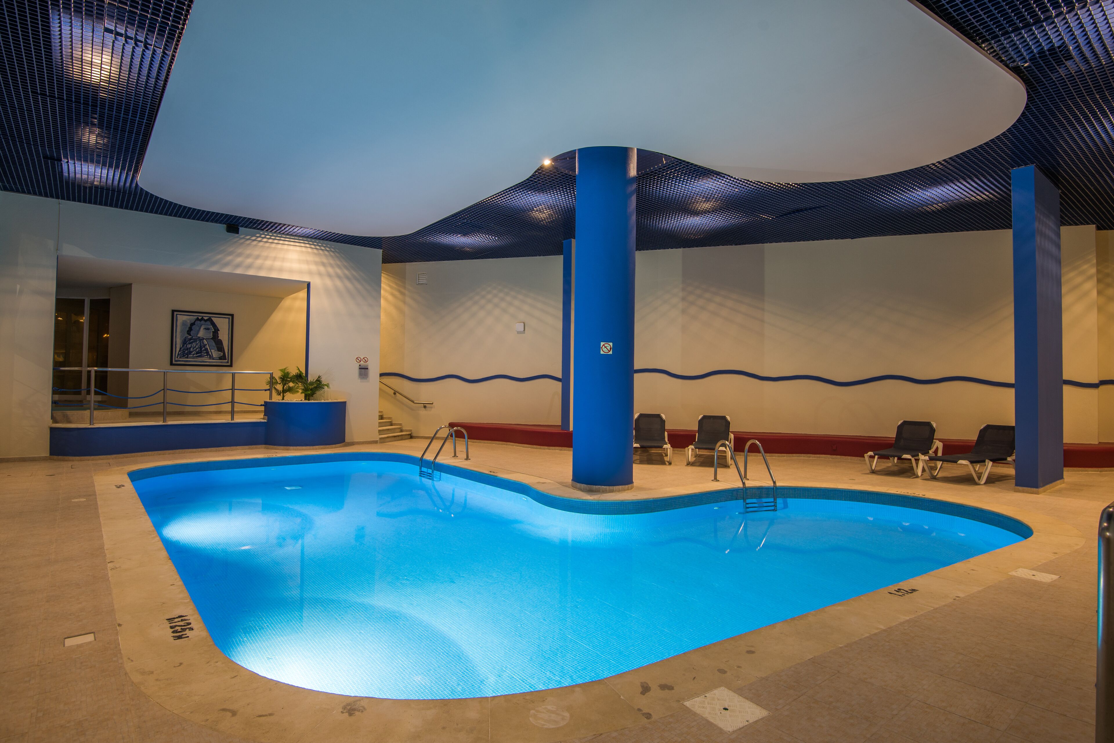 An indoor swimming pool with blue water, surrounded by lounge chairs and a staircase leading to an upper level.