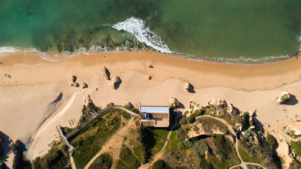 Vista aérea de una playa con un edificio, aguas cristalinas y formaciones rocosas.