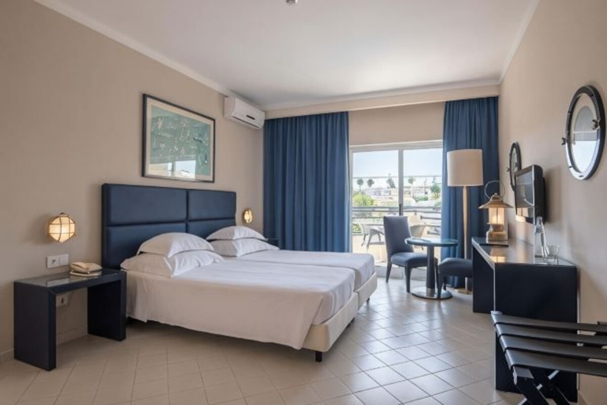A hotel room with a bed, bedside table, lamp, chair, and a view of the outdoors.