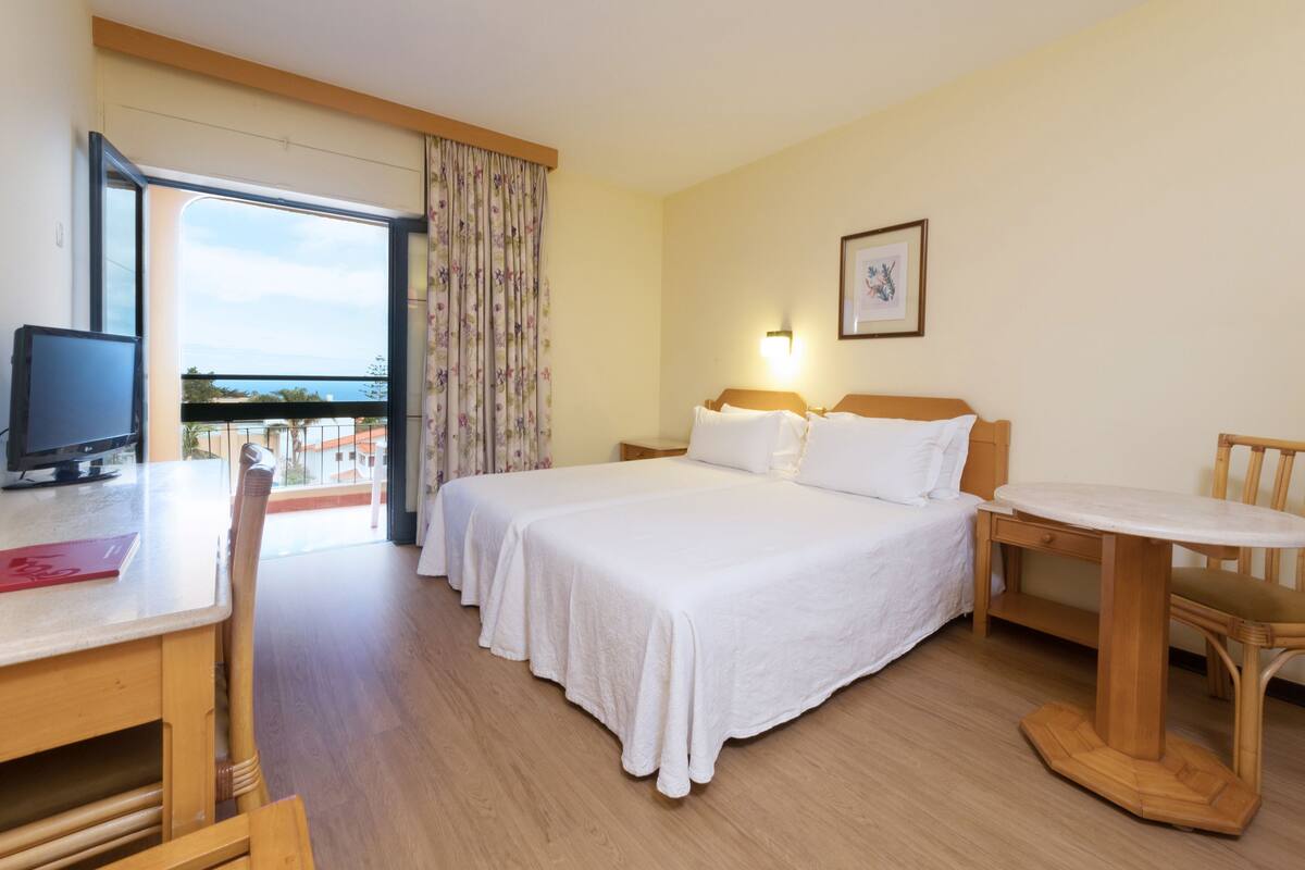 A hotel room with a bed, a desk with a computer, a chair, a small table, and a balcony with a view.
