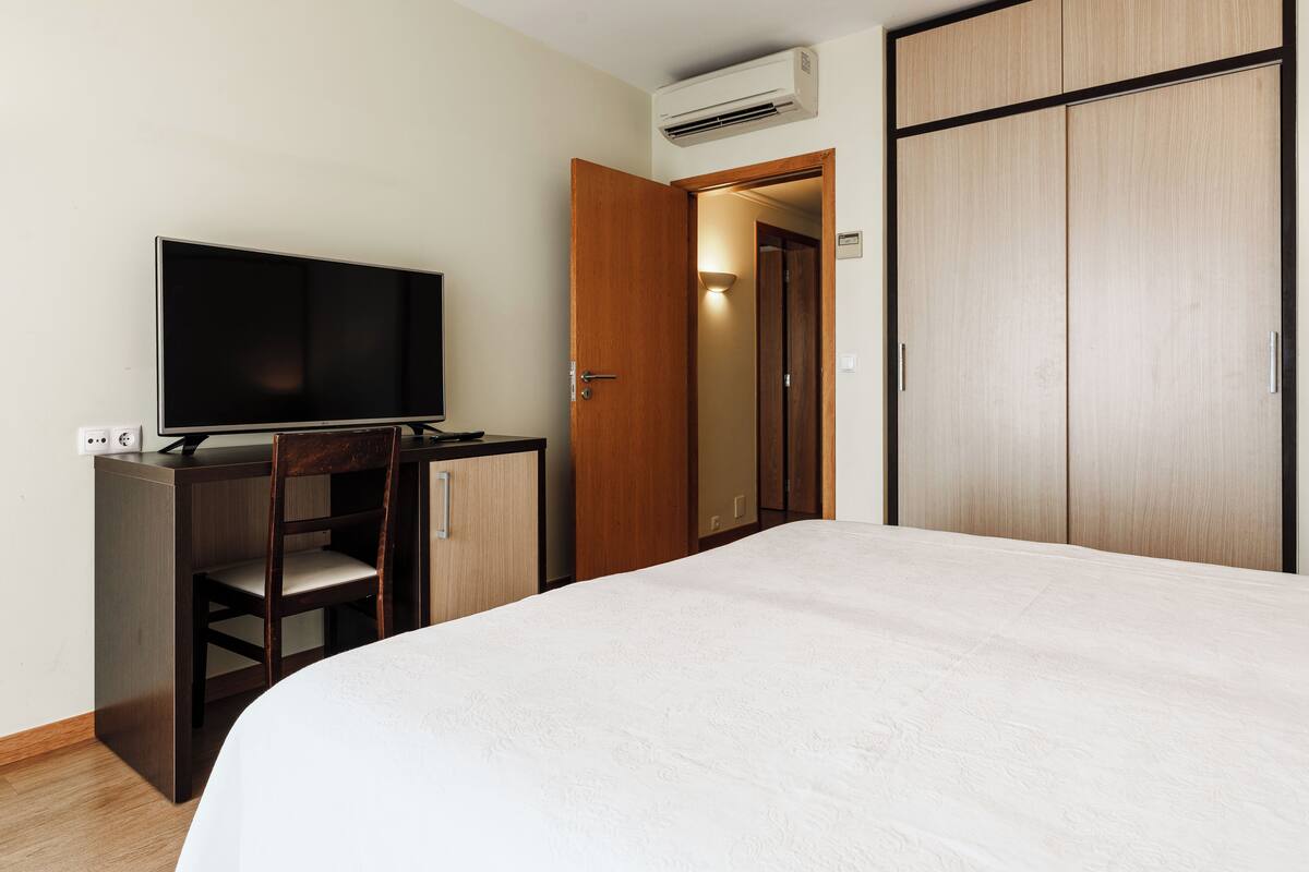 A bedroom with a bed, a TV on a desk, a wardrobe, and an air conditioning unit.