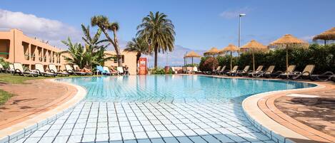 A swimming pool with lounge chairs and palm trees, surrounded by a building with a tiled walkway.