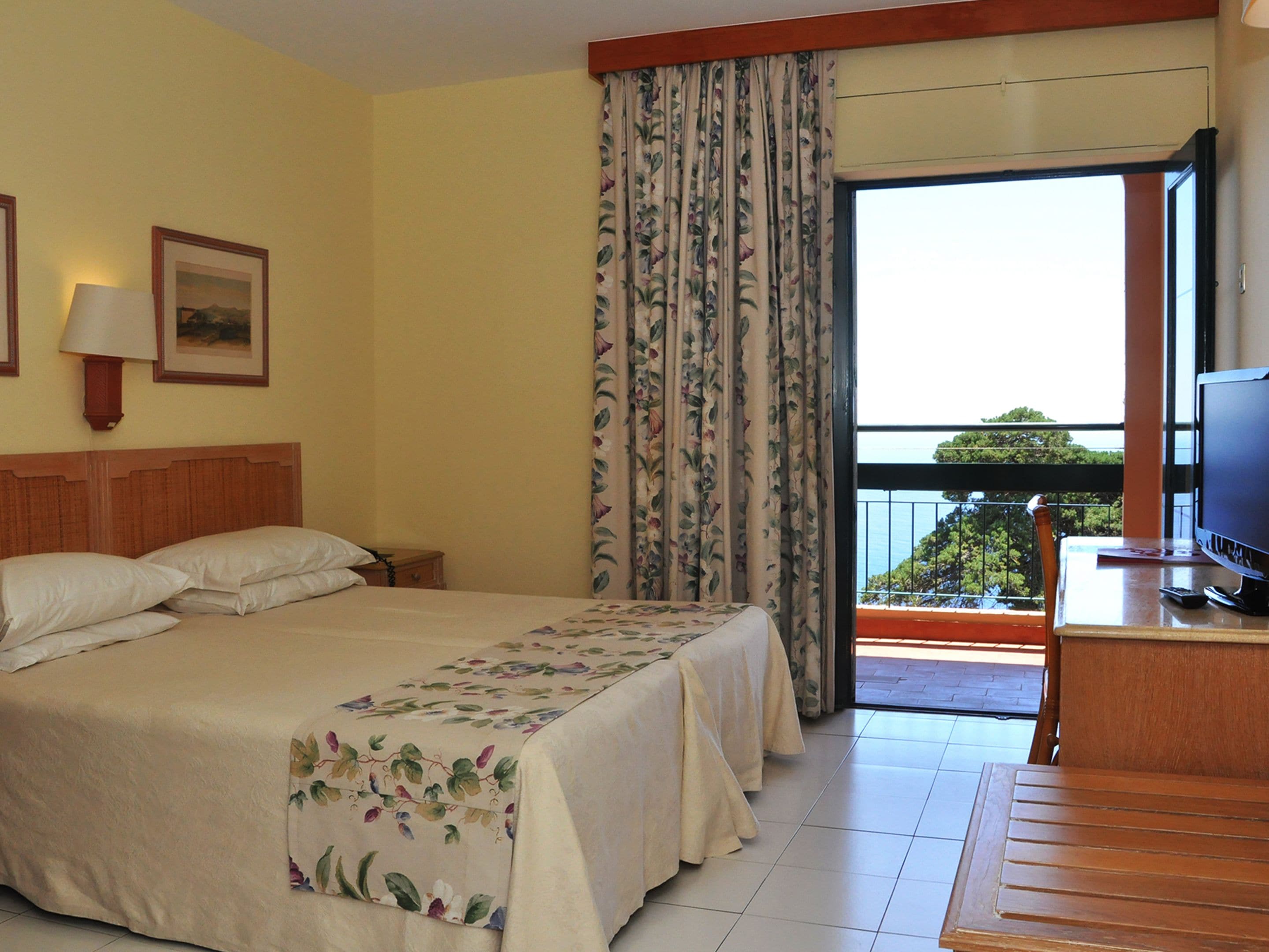 A hotel room with a bed, a desk with a television, a window with curtains, and a picture on the wall.