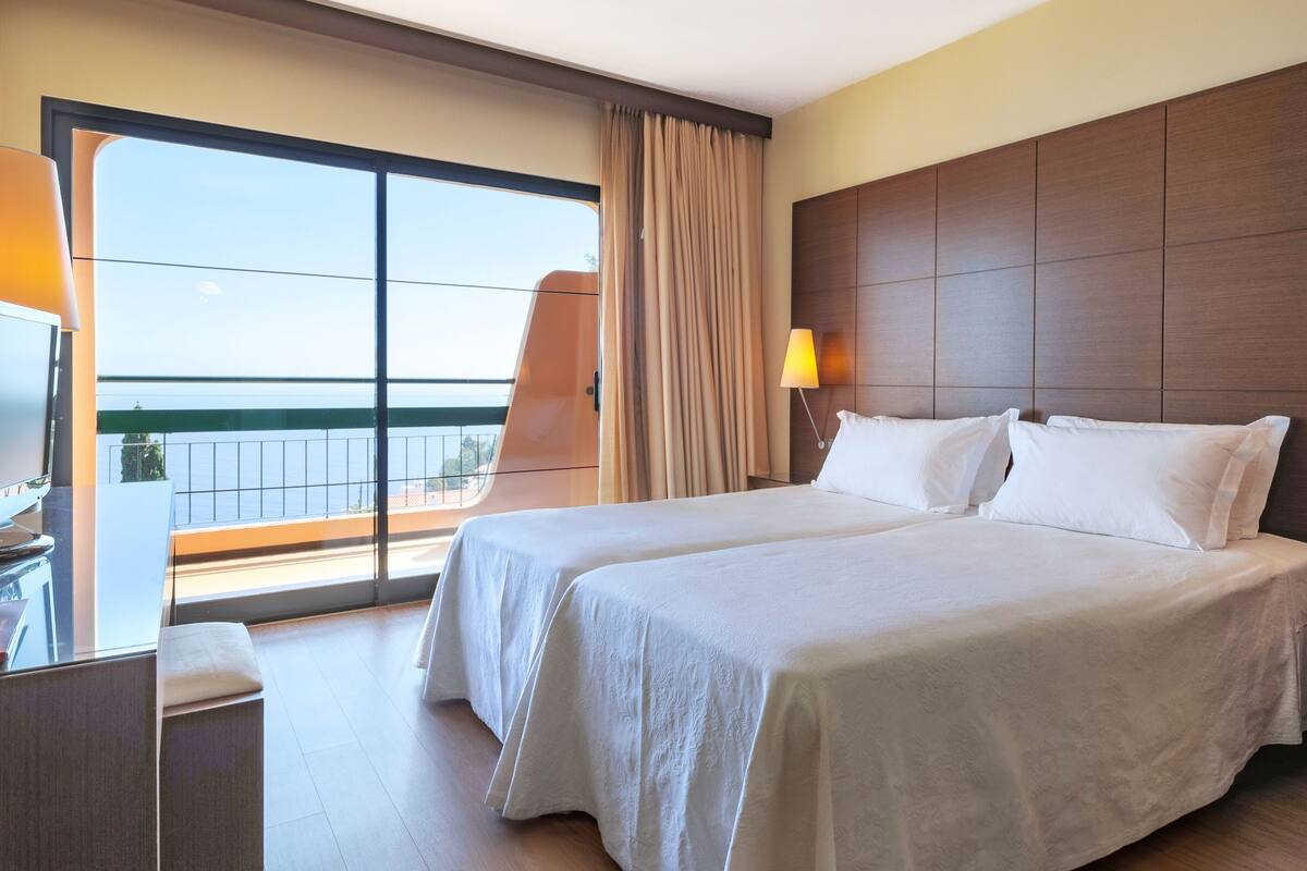 A hotel room with two beds, a desk, a chair, a lamp, and a view of the sea through large windows.