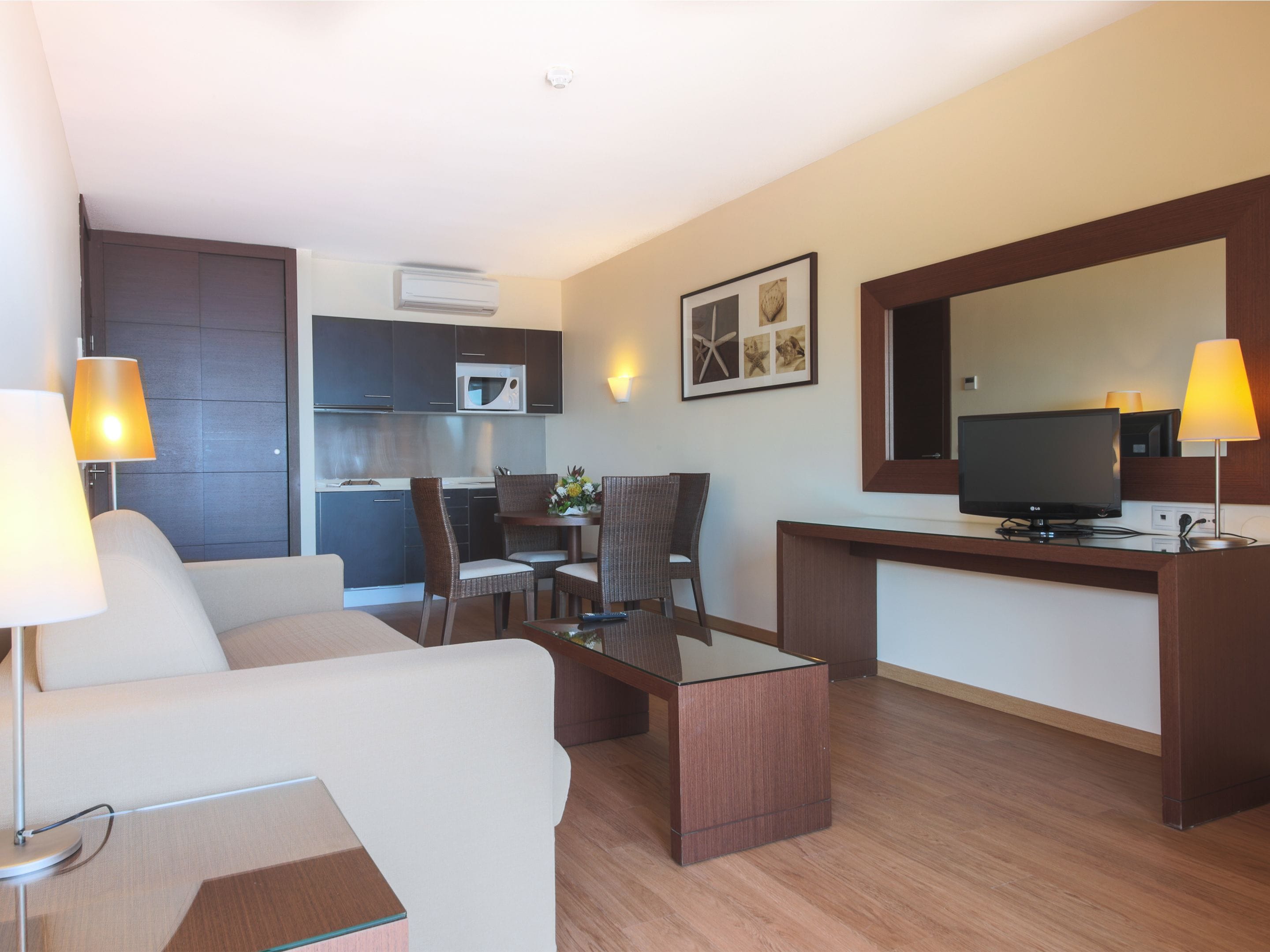 Une chambre d’hôtel comprenant un canapé, une table basse, un téléviseur, un bureau, une chaise, une petite table avec un vase, et un coin cuisine équipé d’un four à micro-ondes.