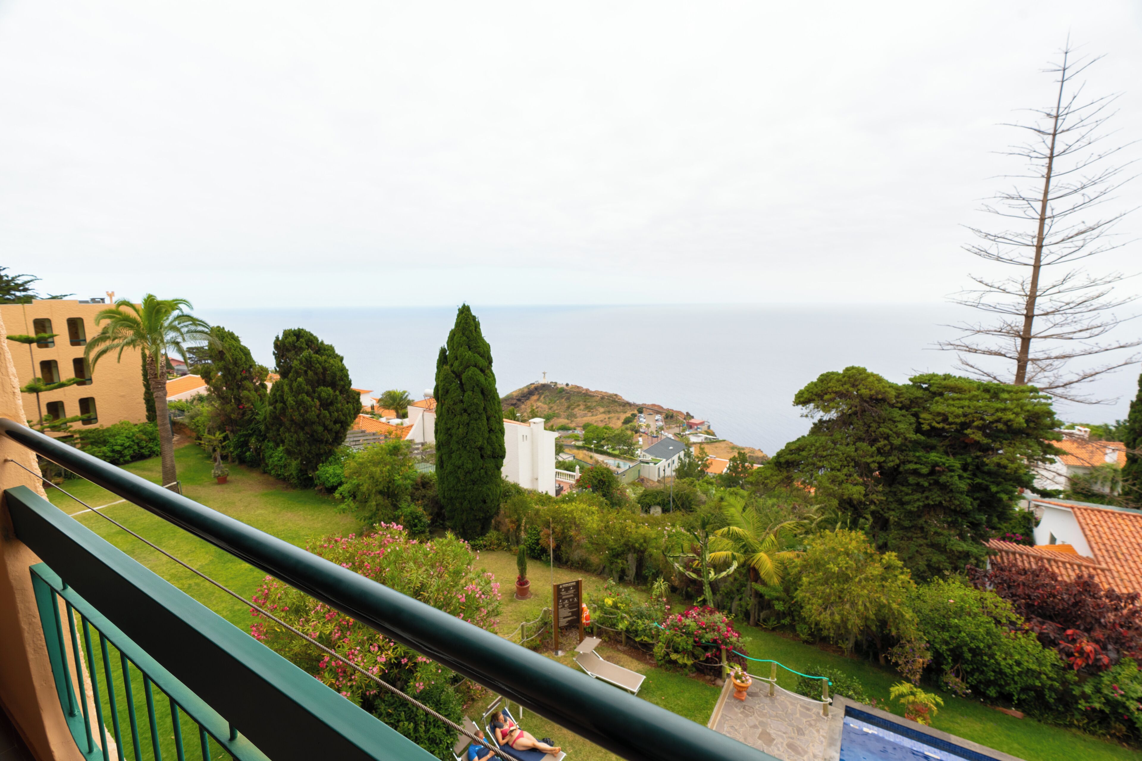 A balcony with a view of a garden, a pool, and the sea.