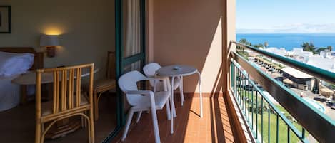 Varanda com uma cama, uma mesa e duas cadeiras. A varanda oferece vista para o mar e edifícios.