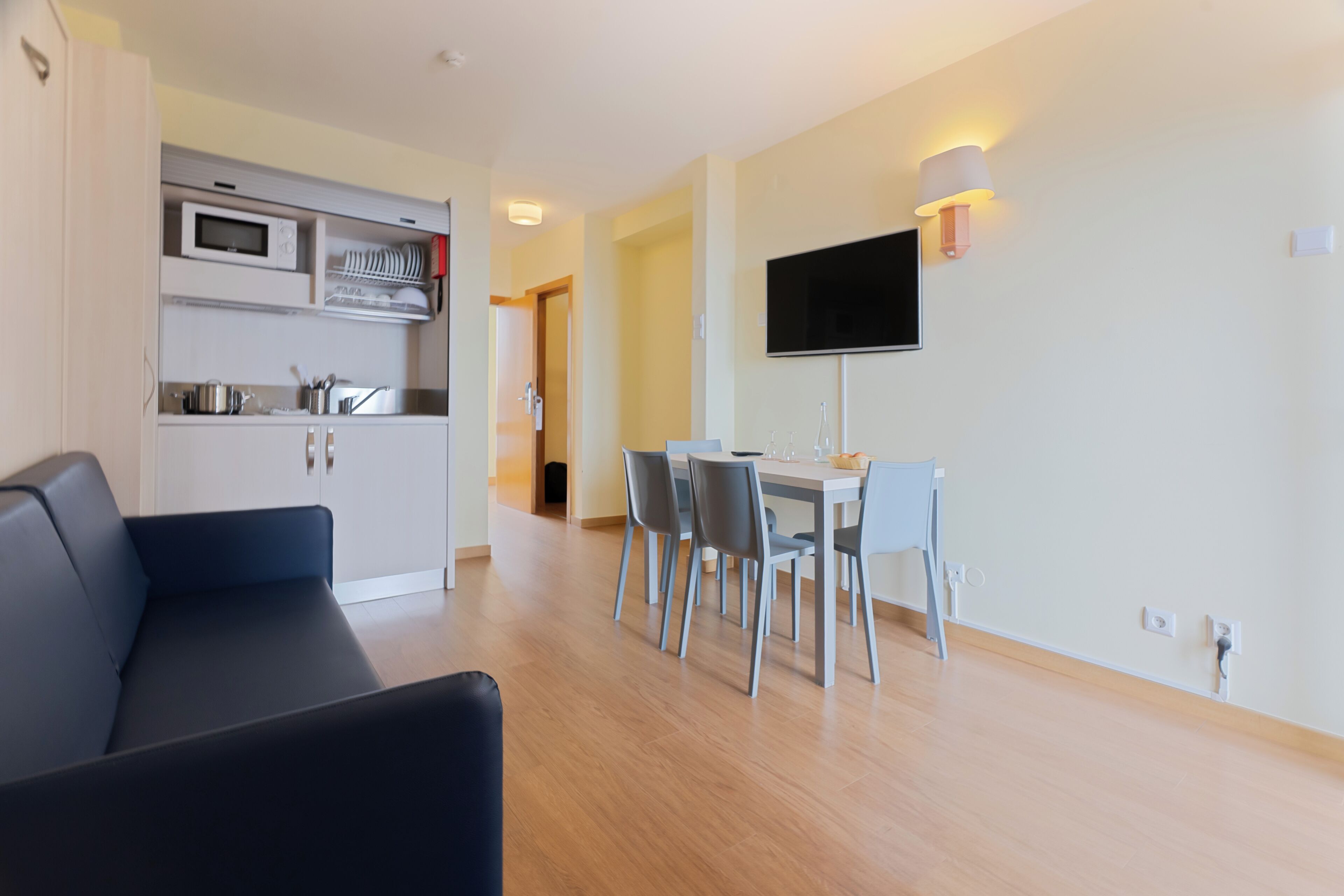 A modern living room with a sofa, a dining area with a table and chairs, a kitchen with appliances, and a TV mounted on the wall.