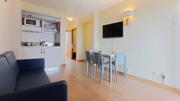 A modern living room with a sofa, a dining area with a table and chairs, a kitchen with appliances, and a TV mounted on the wall.