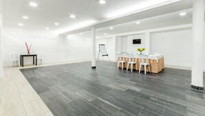 Una habitación amplia con piso de madera oscura, paredes blancas y techo blanco con iluminación empotrada. Se observa una pizarra, una mesa con sillas y una pequeña planta sobre la mesa.
