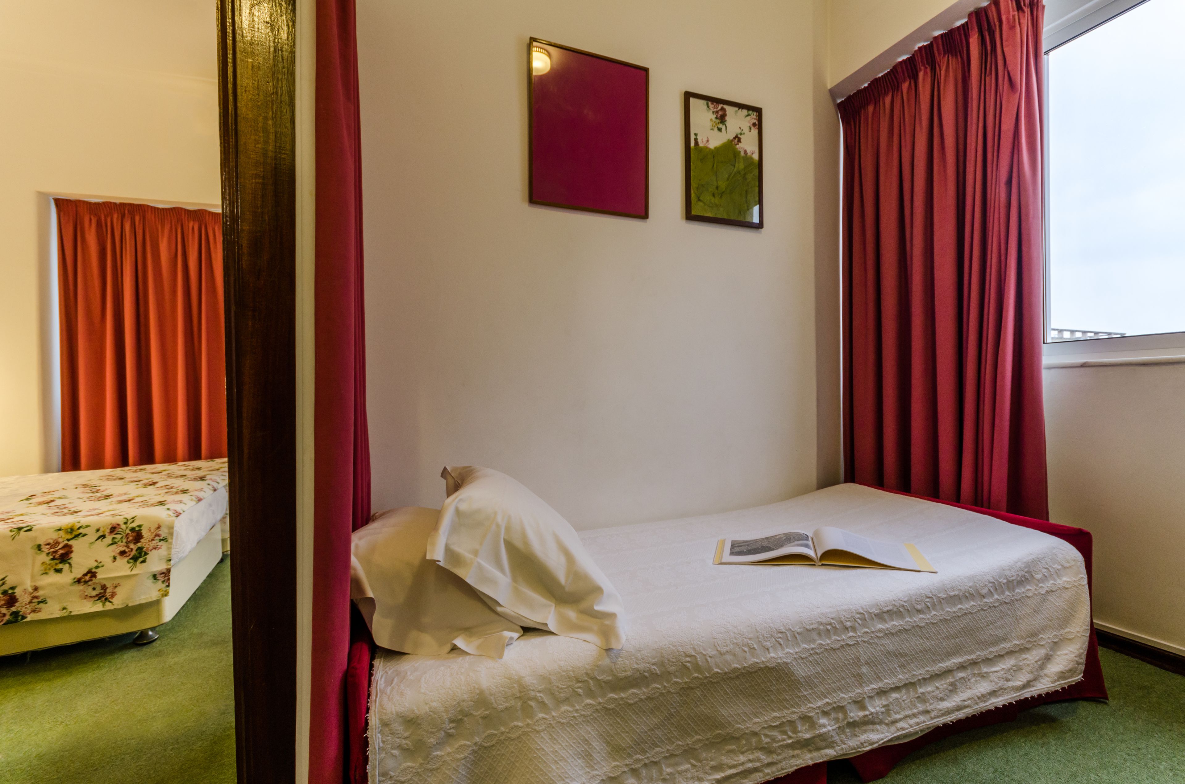 A single bed with a white bedspread and a book on it, a small bedside table, and a window with red curtains.