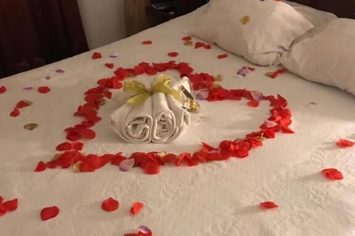 Un lit bien fait, agrémenté d’une composition en forme de cœur faite de pétales de roses et d’une serviette roulée ornée d’un ruban jaune.