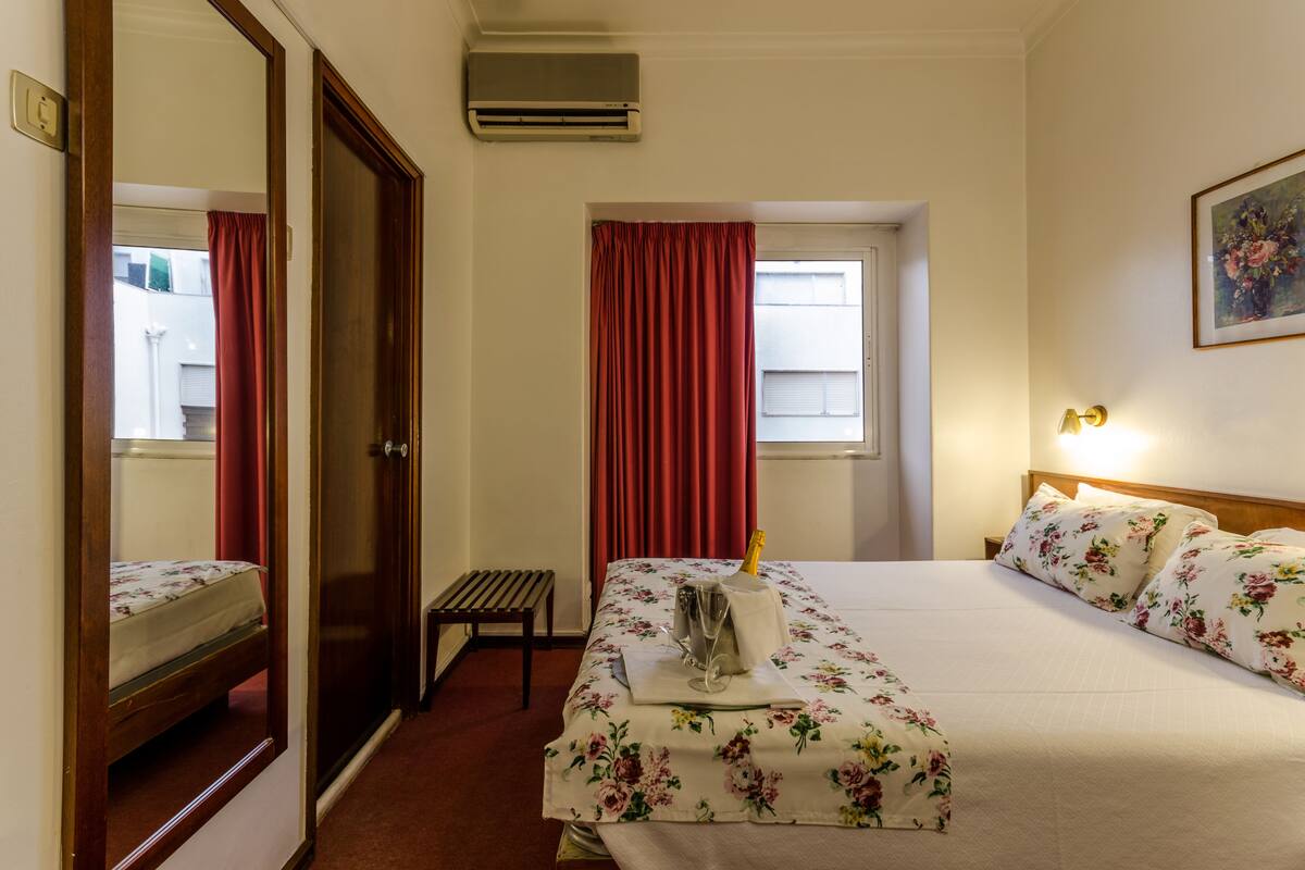 Une chambre d’hôtel avec un lit, une banquette, une fenêtre avec des rideaux rouges et un climatiseur fixé au mur.